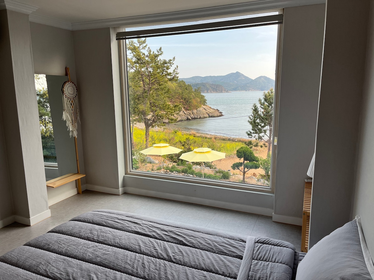 A serene bedroom features a large window framing a scenic coastal view. Soft sunlight illuminates a neatly made spacious bed, while a mirror reflects elements of the peaceful outdoor landscape, including yellow umbrellas and lush greenery.