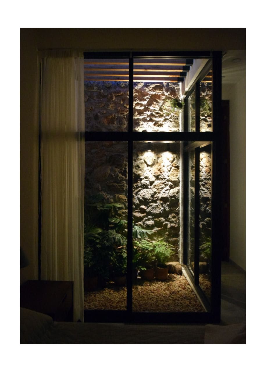 An indoor garden area is visible through large glass doors, featuring natural stone walls and various potted plants. Soft lighting enhances the serene ambiance, while sheer curtains gently frame the view, allowing for a connection with the greenery outside.