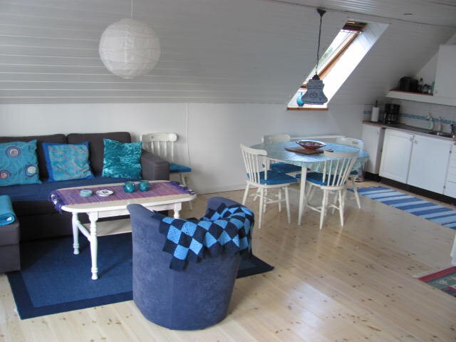 An inviting living space features a comfortable sofa adorned with turquoise and blue cushions. A round dining table sits beside a kitchenette, with light streaming through a slanted window. A cozy chair and a patterned blanket complement the warm wooden floors.