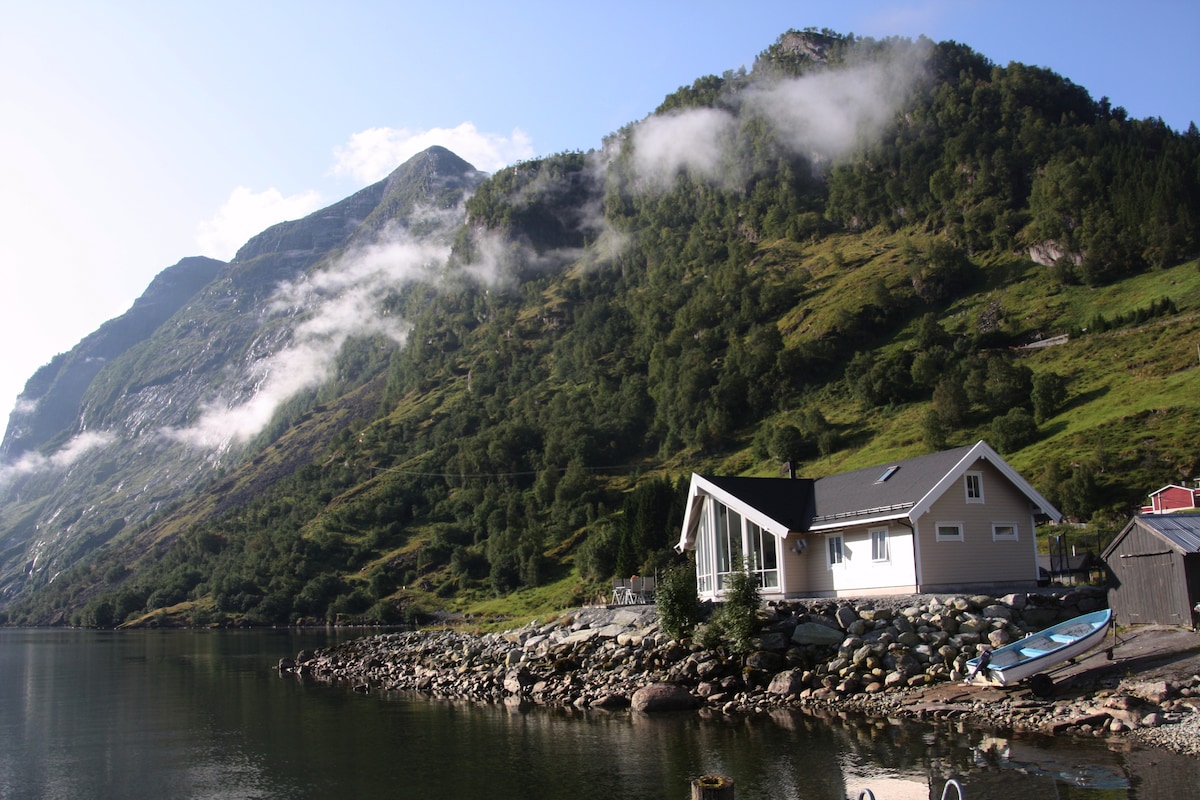 A cabin is situated along the shore of a calm lake, with high mountains rising majestically in the background. Lush greenery covers the mountainsides, while soft clouds drift above, creating a serene natural setting. A small dock and boats are partially visible at the water's edge.