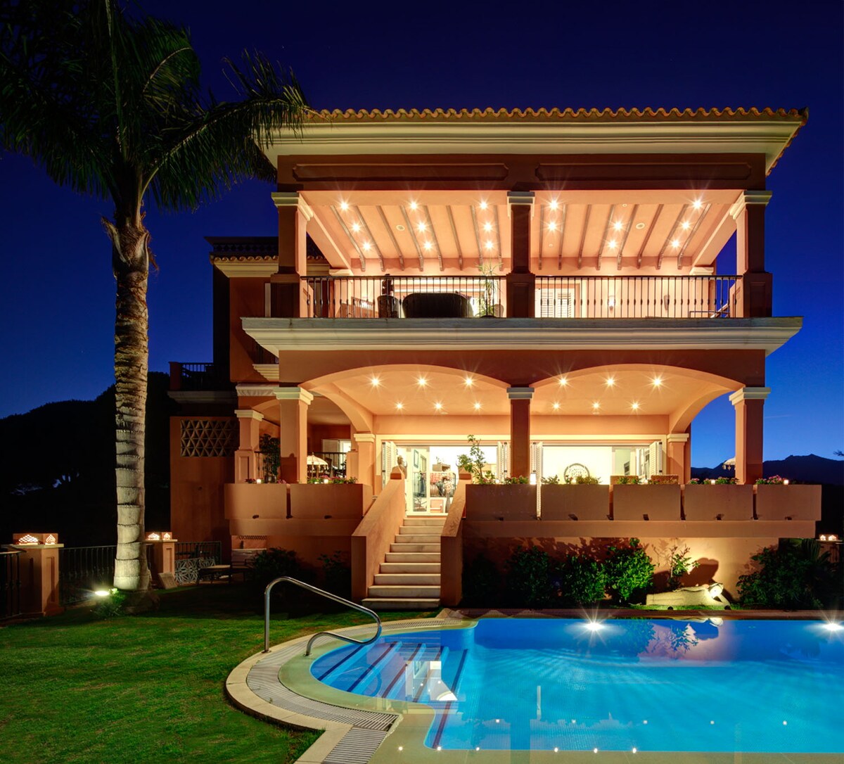 A three-story villa is illuminated against the evening sky, with multiple balconies showcasing elegant outdoor seating. A swimming pool with gentle lighting reflects the structure's warm colors, while palm trees enhance the serene outdoor setting.