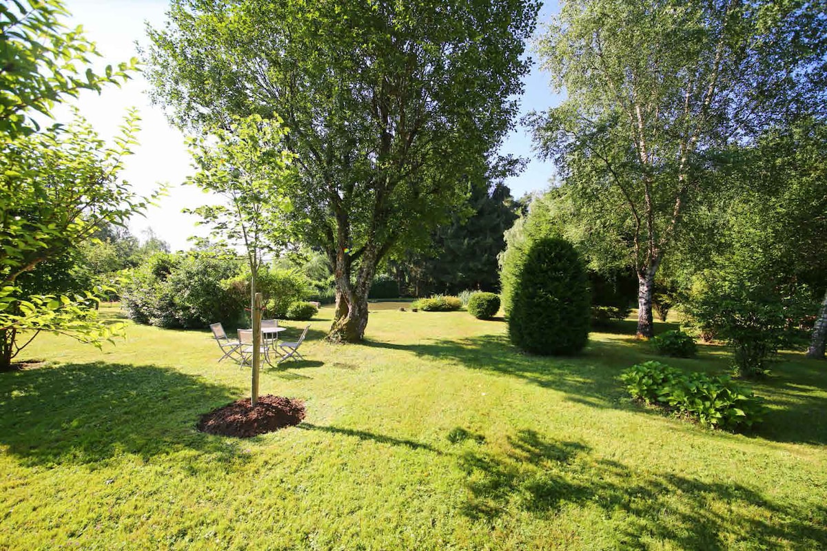 A spacious green garden is featured, showcasing a variety of trees and shrubs. A small table with two chairs is positioned on the grass, providing a peaceful area for outdoor relaxation. Sunlight creates gentle shadows across the well-maintained lawn.