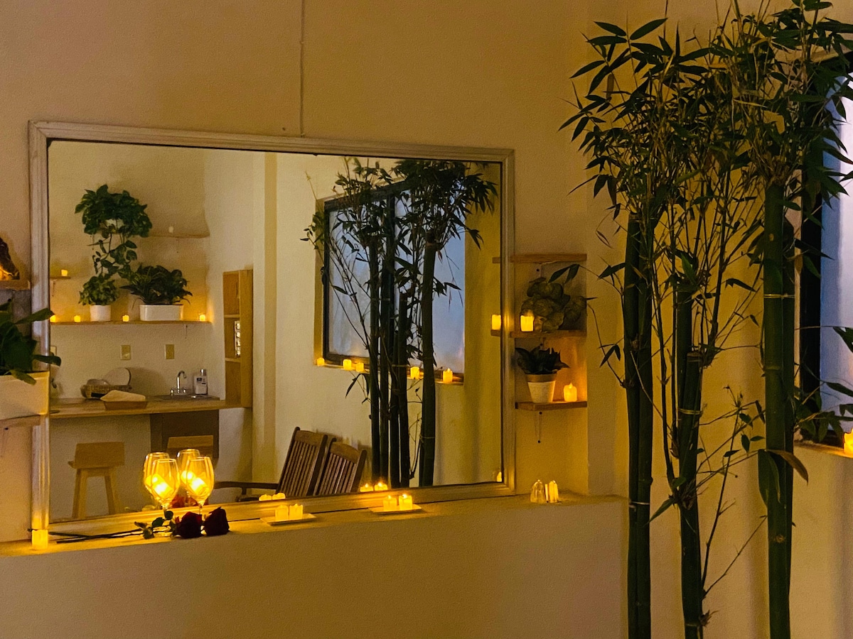A spacious mirror reflects a serene interior featuring tall bamboo plants on either side. Soft candlelight glows warmly throughout the room, while shelves hold decorative plants. A kitchen area is subtly visible in the background, enhancing the cozy atmosphere.