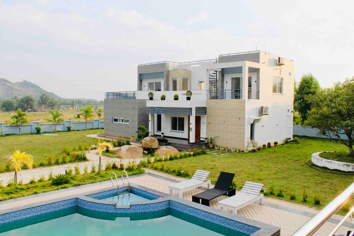 A modern two-story house is shown, featuring large windows and a balcony. The well-maintained lawn surrounds a rectangular swimming pool, accompanied by lounge chairs. The scenic backdrop includes gentle hills, offering a serene outdoor environment.