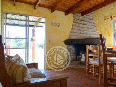 A cozy living space features a stone fireplace, wooden ceiling beams, and a rustic dining area. Light filters in through large glass doors, providing views of the surrounding trees. Comfortable seating is arranged near the warm décor, inviting relaxation.