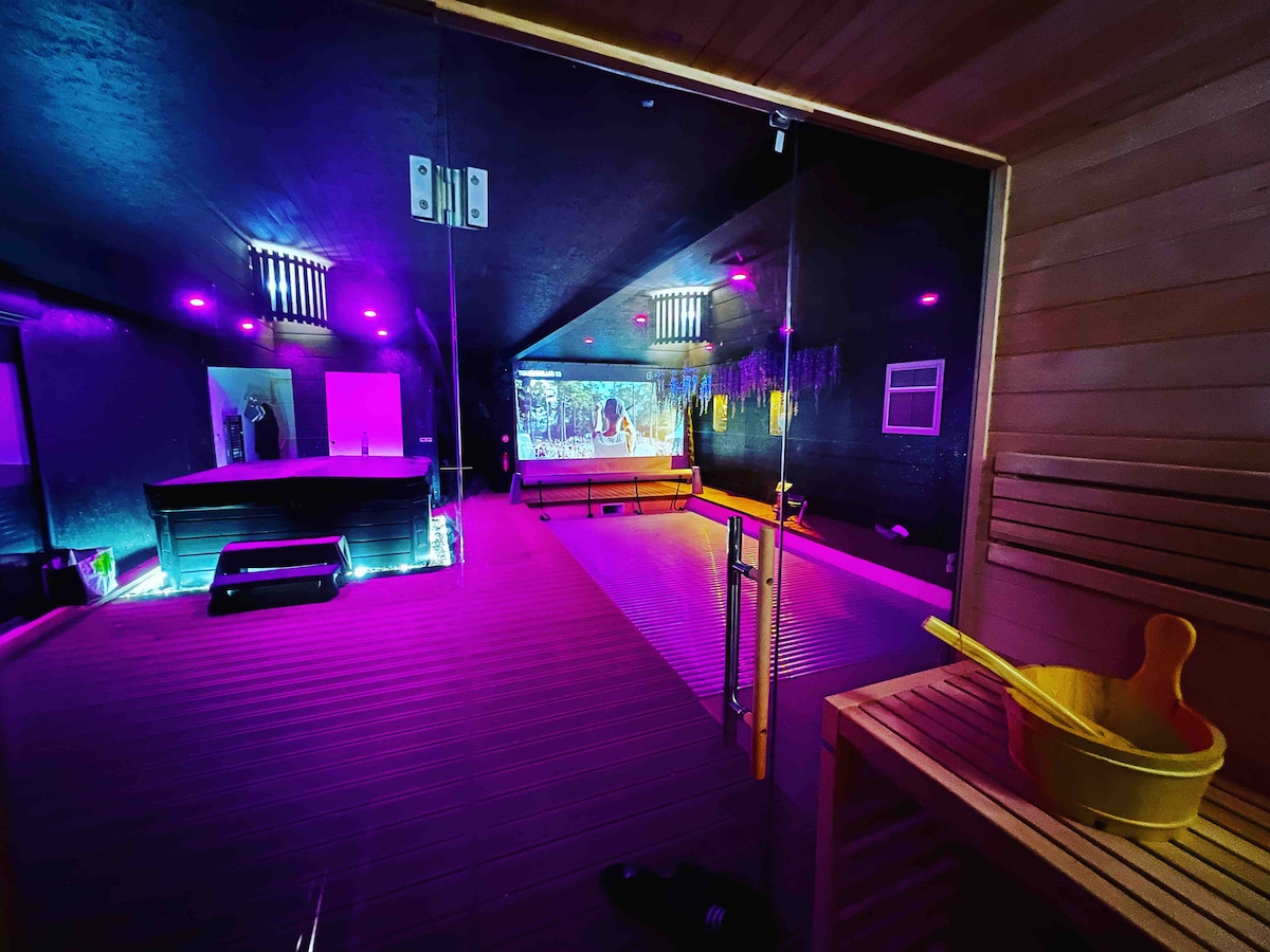 The image portrays a private spa area featuring warm lighting in shades of pink and purple. A wooden sauna bench is visible, alongside a yellow scoop. A relaxation zone is shown with a bar, enhancing the cozy ambiance for guests.