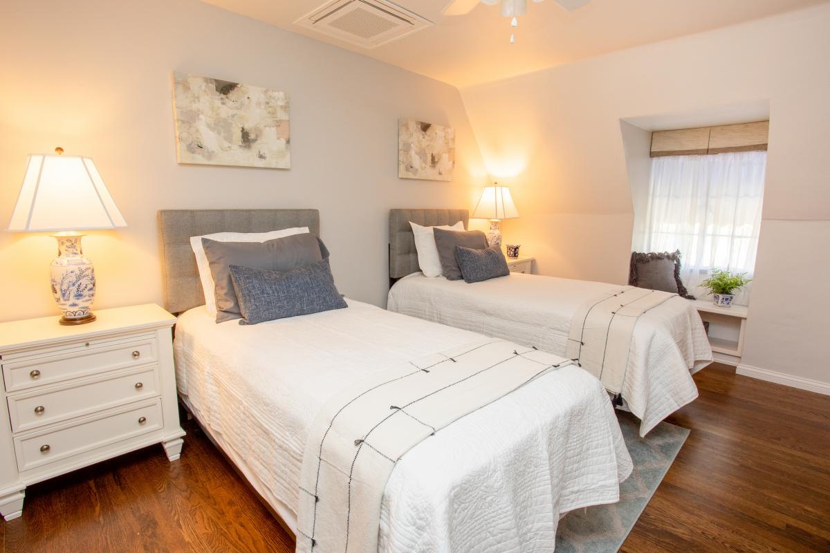 A serene sleeping area features two twin-sized beds with white bedding and grey accent pillows. Each bed is flanked by stylish bedside lamps. Natural light filters through a window, accompanied by light white curtains. A cozy nook is visible, with a small plant on a shelf.