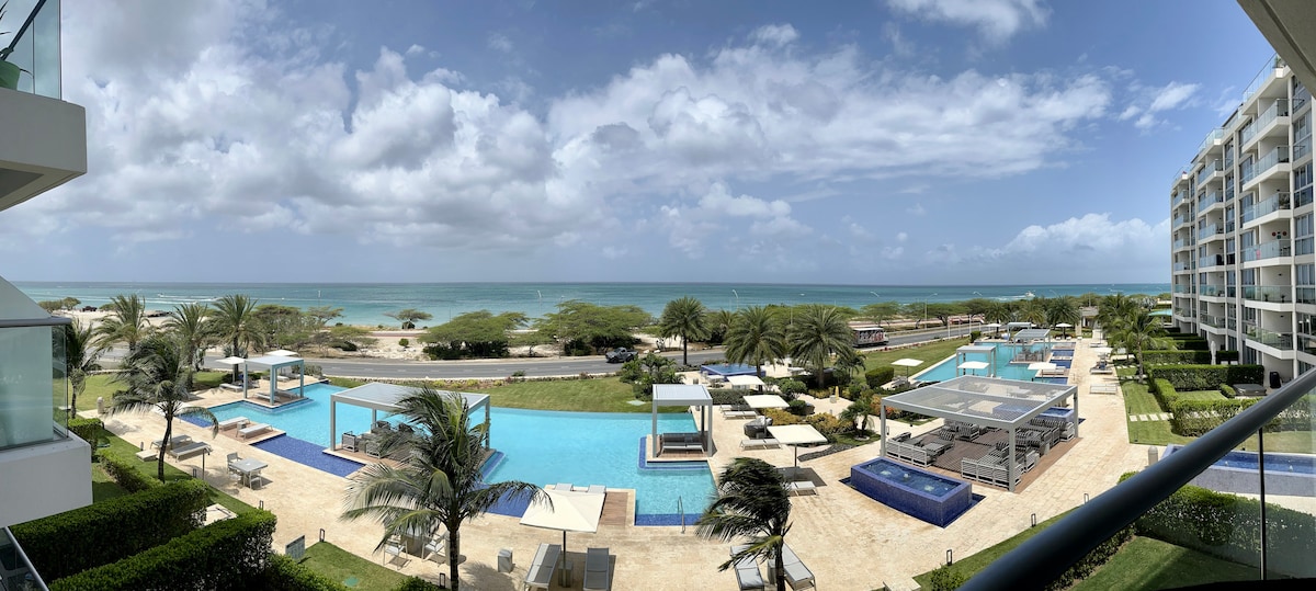 A panoramic view showcases the resort-style outdoor space, featuring two swimming pools and a jacuzzi. Lounge areas with shaded cabanas surround the pools, with palm trees and ocean in the background. The vibrant coastline is visible, enhancing the overall atmosphere.