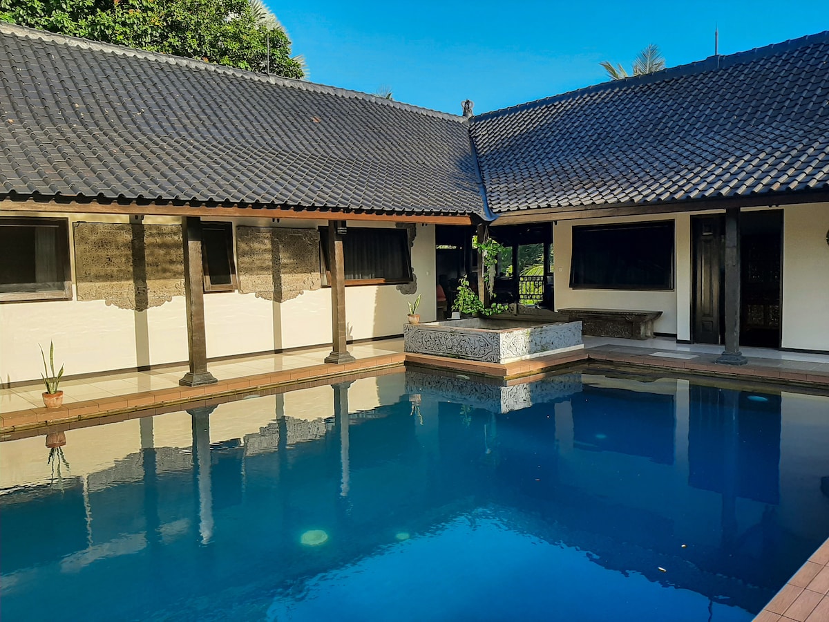A serene pool area is framed by traditional architecture with a thatched roof. Clear water reflects the blue sky and surrounding greenery. The space is complemented by a stone feature at the pool's edge, inviting relaxation and enjoyment of the outdoors.