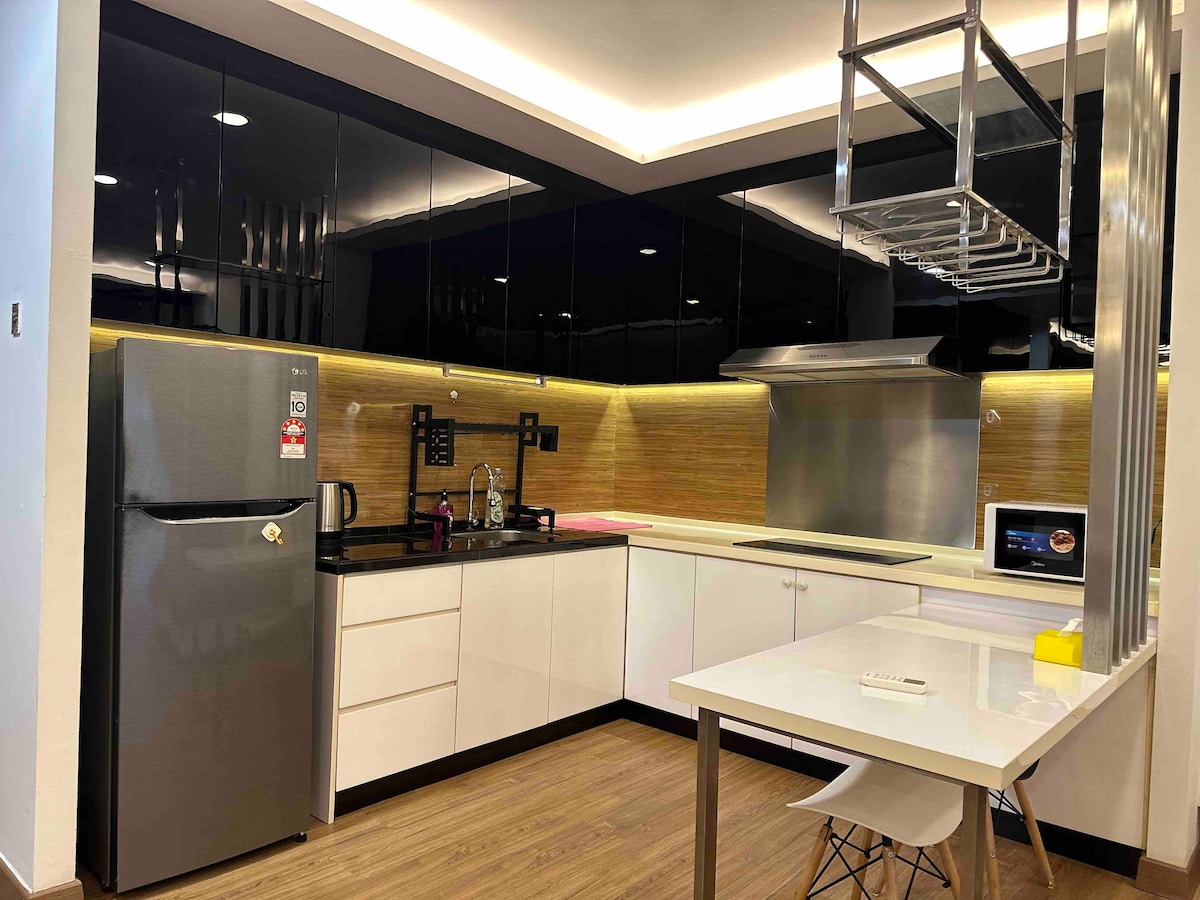 A modern kitchenette is displayed, featuring sleek black cabinetry and a wooden backsplash. Included are a refrigerator, kettle, stovetop, and oven. A small dining table with a minimalist design is positioned beside the kitchen, providing an inviting space for meals.
