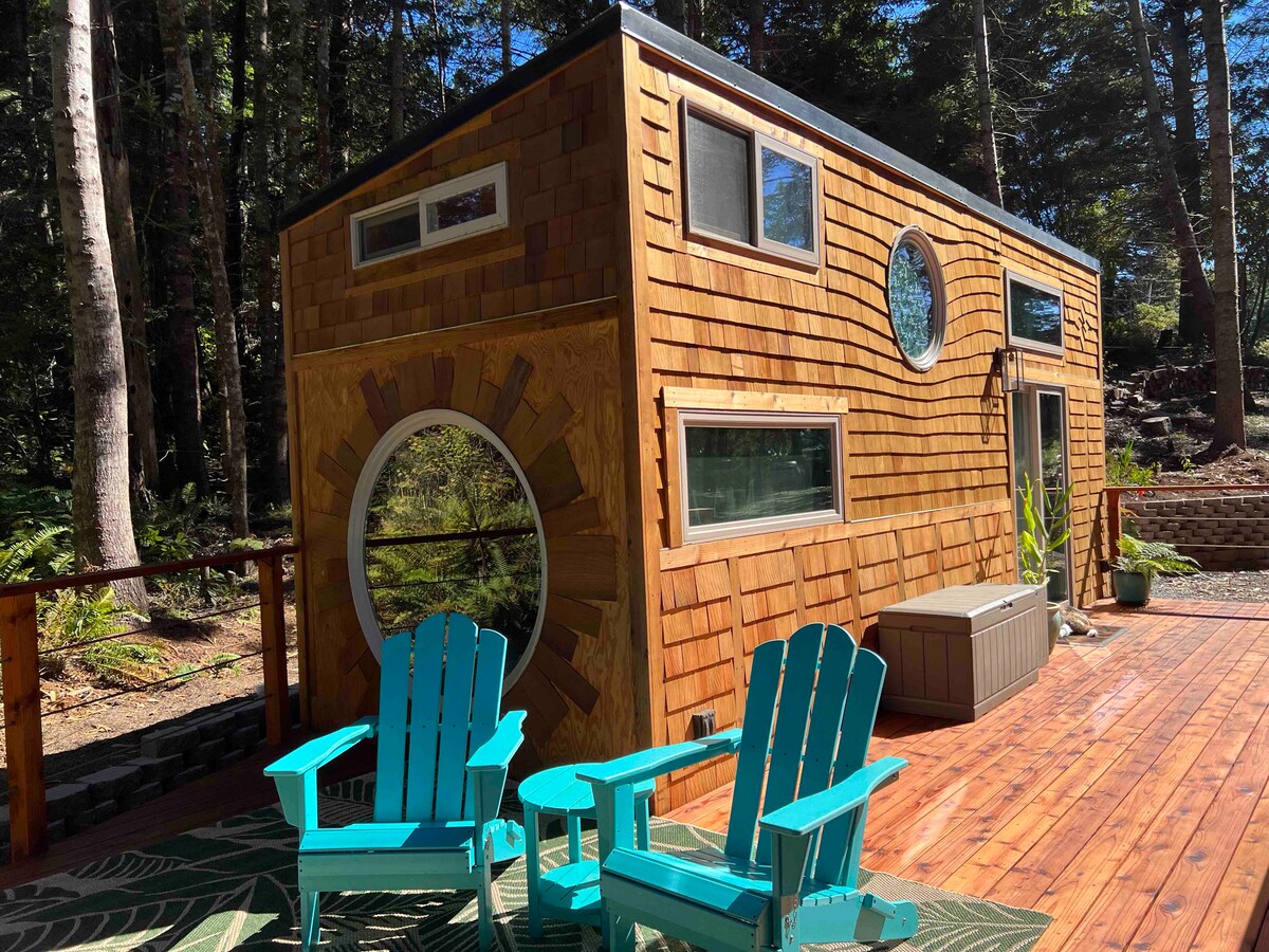 A charming cabin exterior features a mix of circular and rectangular windows, allowing natural light to fill the interior. A spacious deck is complemented by two turquoise Adirondack chairs, providing a serene spot to enjoy the surrounding forest. Wood accents enhance the cabin's rustic appeal.
