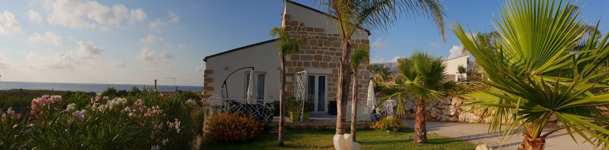 A quaint accommodation is surrounded by lush greenery and colorful flowers, providing a peaceful atmosphere. The structure features a light facade with stone accents and large glass doors that open towards a scenic view of the coastline.