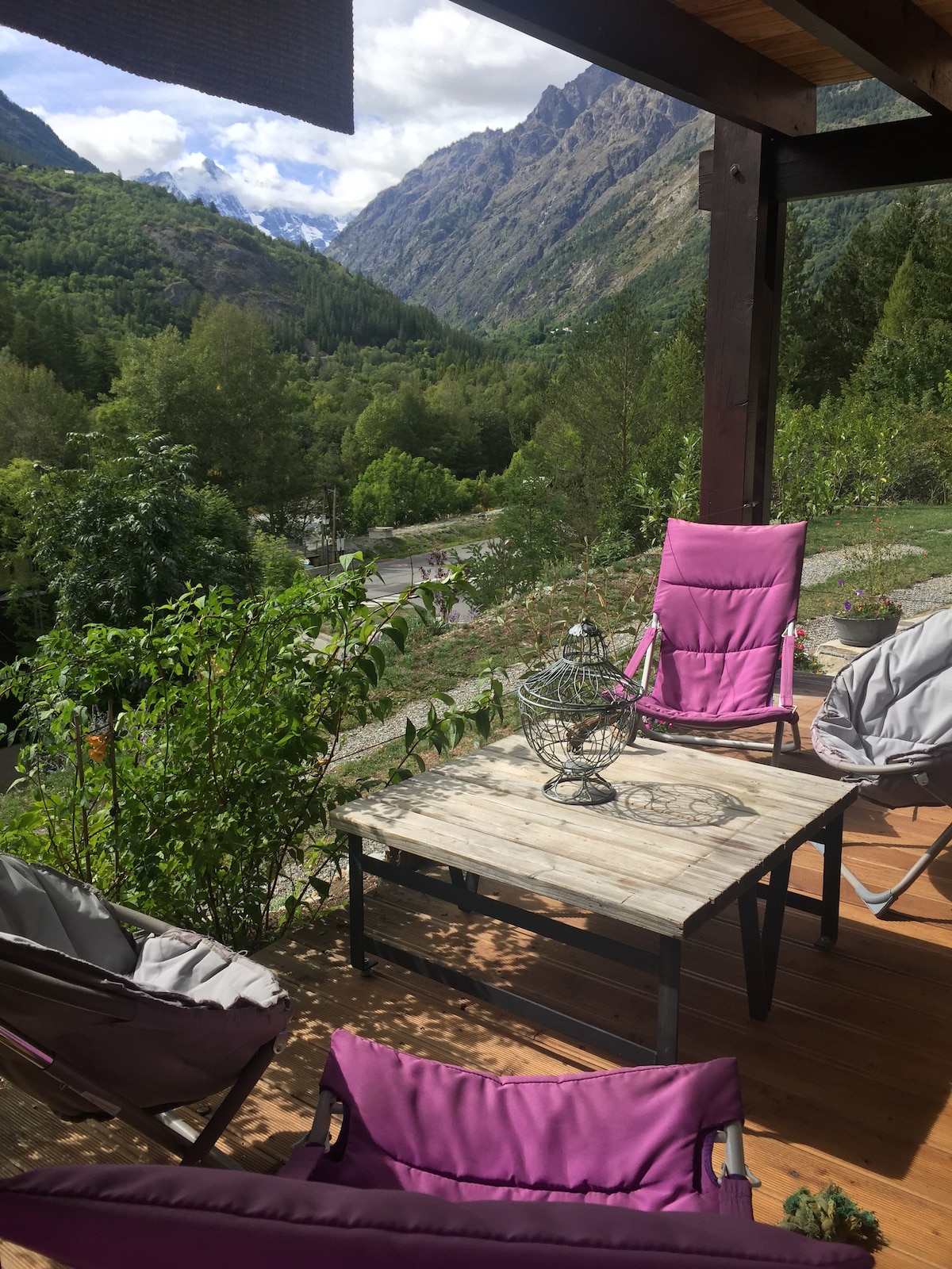 A covered terrace features two purple lounge chairs and a wooden coffee table. Surrounding greenery provides a natural ambiance, with mountainous scenery in the background. Light filters through the leaves above, creating a serene outdoor space for relaxation.