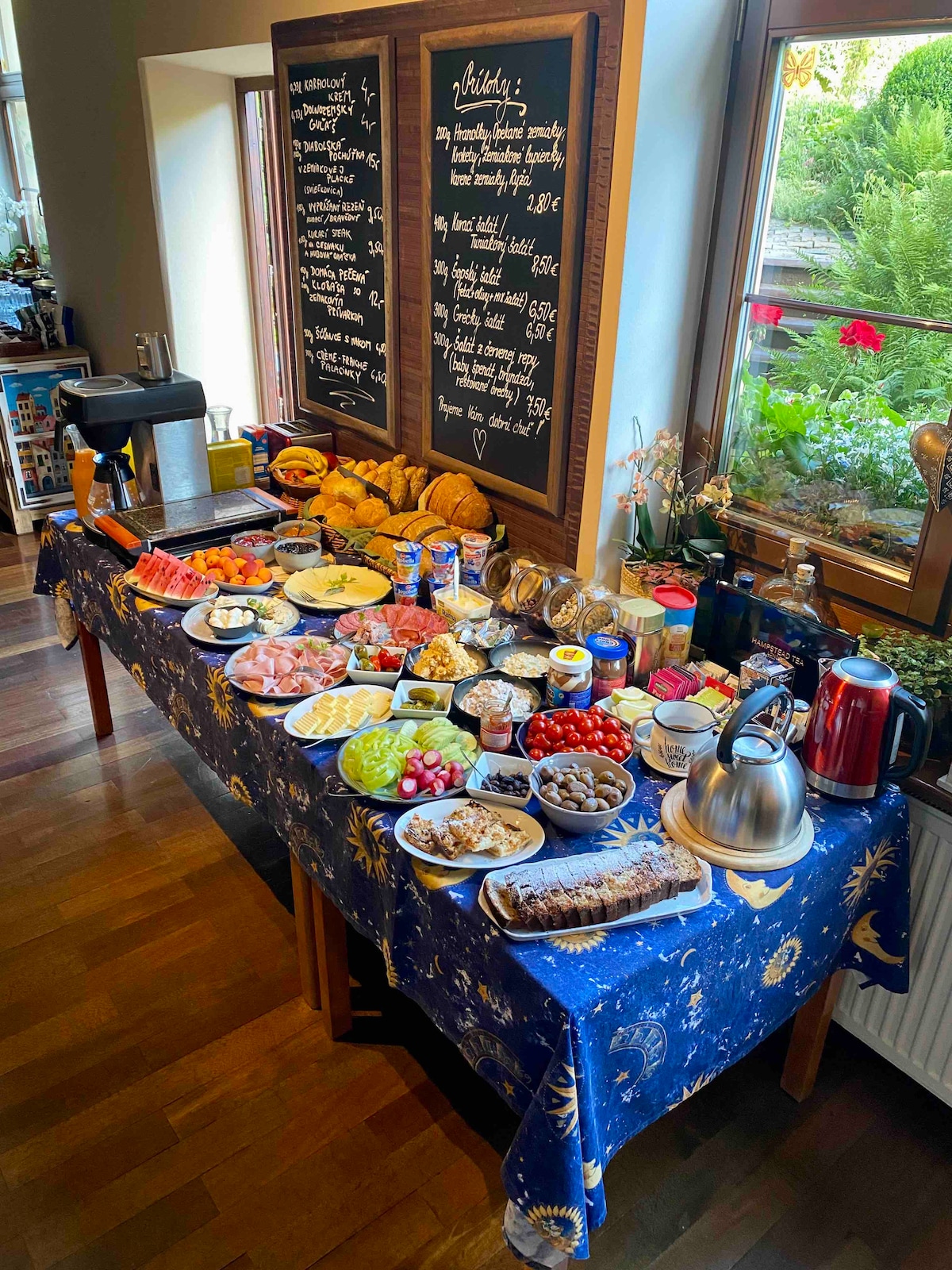 A breakfast spread is set on a table draped in a blue tablecloth with celestial patterns. Various food items are arranged including fresh fruits, meats, bread, and dairy products. Two chalkboards list menu items, while greenery is visible through the windows, enhancing the inviting scene.