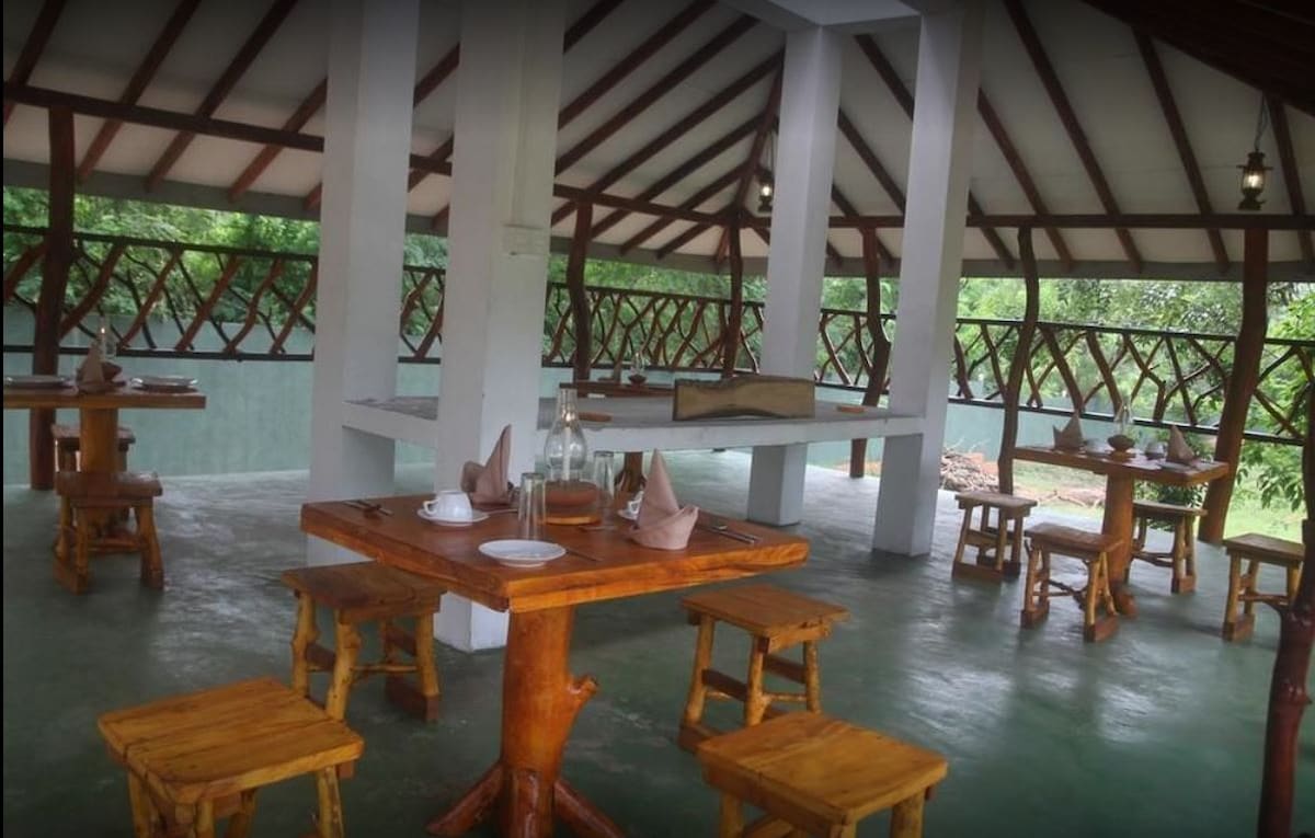 An open dining area features wooden tables and benches arranged under a vaulted roof. Natural light filters in through large windows, providing views of the surrounding greenery. Simple table settings include plates and folded napkins, enhancing the relaxed atmosphere of the space.