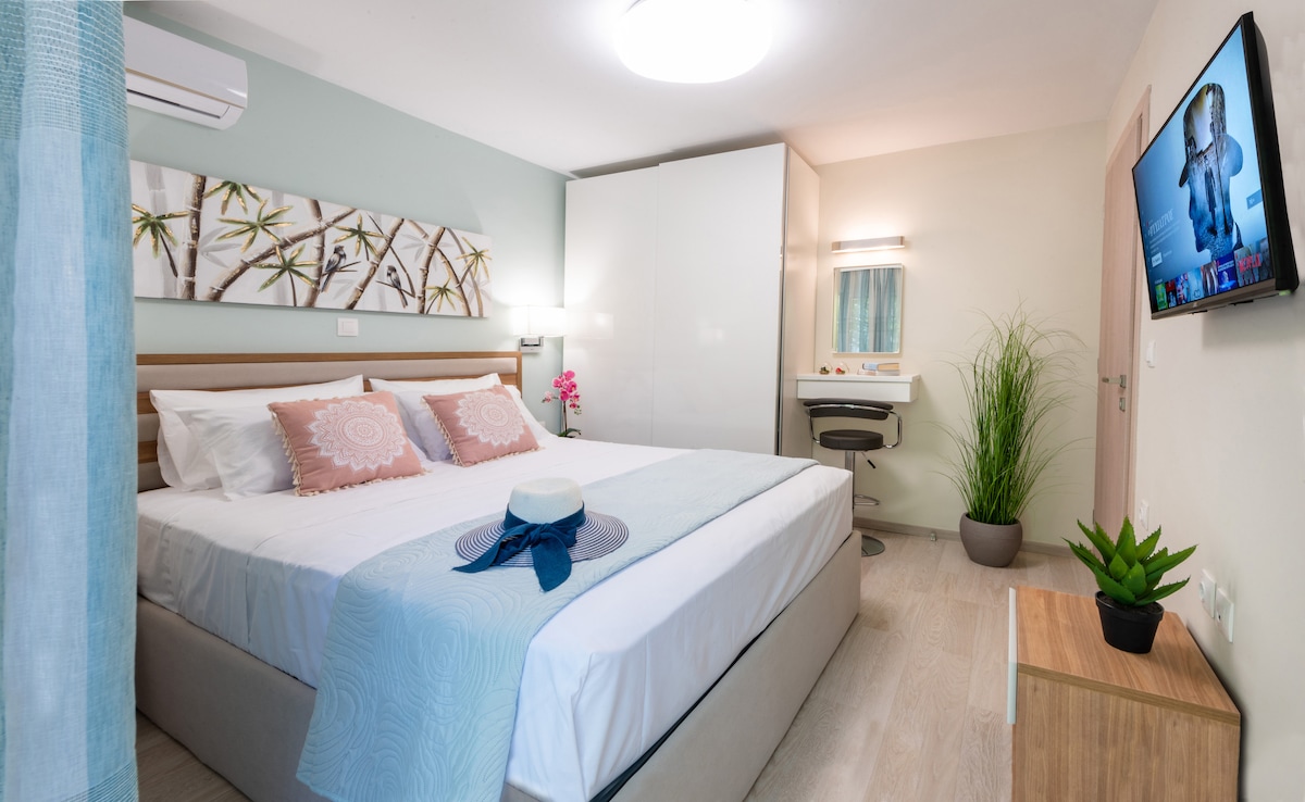 A well-lit bedroom features a spacious bed adorned with decorative pillows and a soft blanket. A modern flat-screen TV is mounted on the wall. Green accents are provided by a potted plant, while a study area with a mirror and chair is visible in the corner.