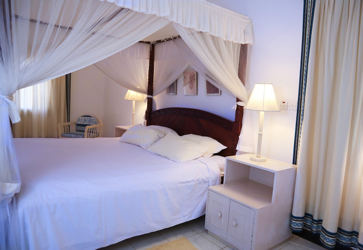 A spacious bedroom features a four-poster bed with a canopy, adorned in white linens. Soft lighting is provided by two bedside lamps beside a wooden nightstand. Light curtains frame a window, allowing natural light to fill the room, which includes a comfortable seating area.
