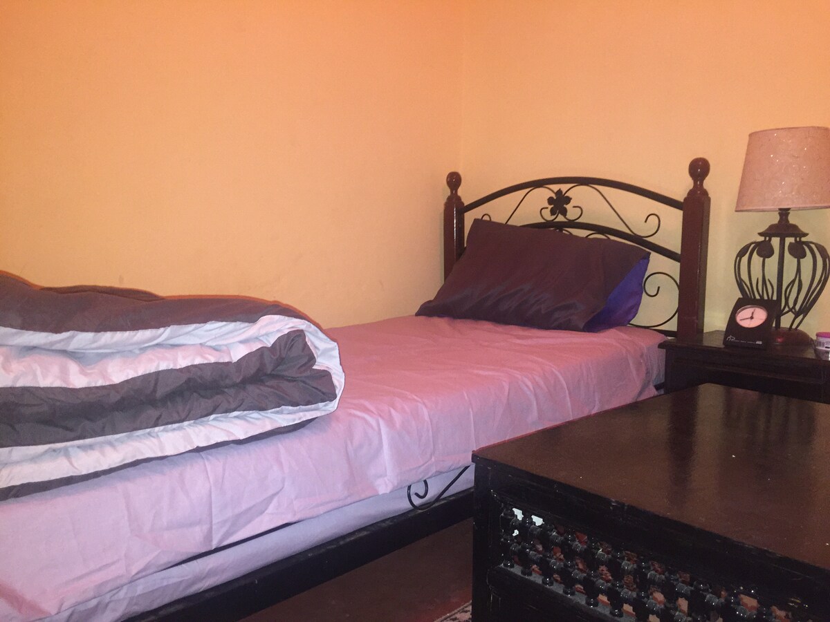 A single bed with a dark iron frame is positioned against a warm-toned wall. A light purple bedsheet covers the mattress, and a neatly folded blanket rests beside it. A small bedside table holds a lamp and a clock, complementing the cozy atmosphere.