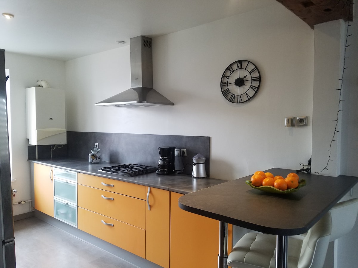 A modern kitchen features a combination of orange and gray cabinetry. The stainless steel appliances include a gas stove and coffee maker. A clock is mounted on the wall, while a small table holds a bowl of oranges. Natural light fills the space through a window.