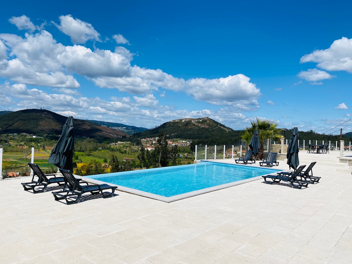 An infinity pool is surrounded by lounge chairs with umbrella shades, offering views of the mountains and lush landscape. The bright blue sky is dotted with clouds, complementing the serene atmosphere of the outdoor space.