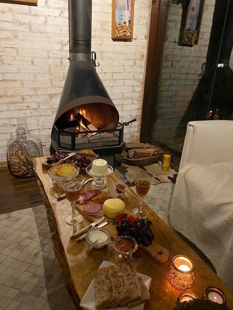 A rustic wooden table is set with a variety of cheeses, cured meats, and fruits. A glowing fireplace is visible in the background, adding warmth to the inviting space. Candles and decorative jars create a cozy ambiance, enhancing the overall setting.
