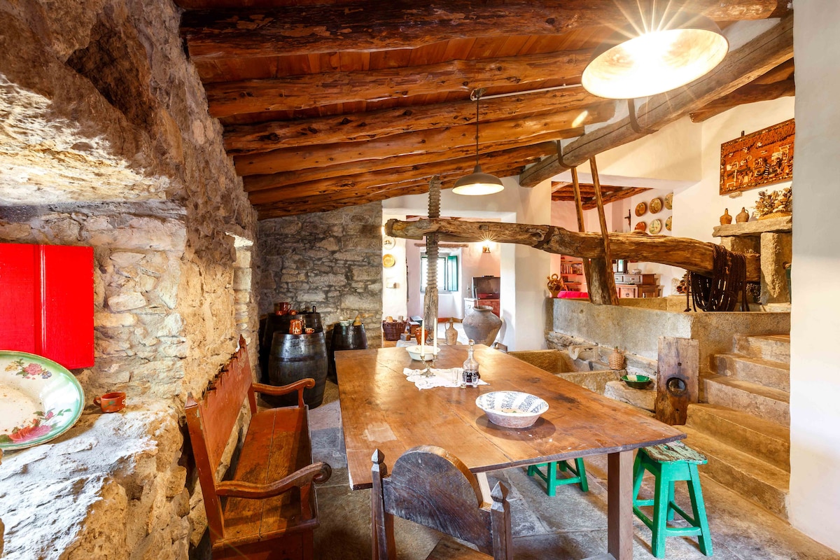 A rustic dining area features a large wooden table surrounded by various colored chairs. Stone walls and wooden beams create a charming ambiance. A mix of decorative items and pottery adorns the space, while natural light filters through a window, enhancing the cozy atmosphere.
