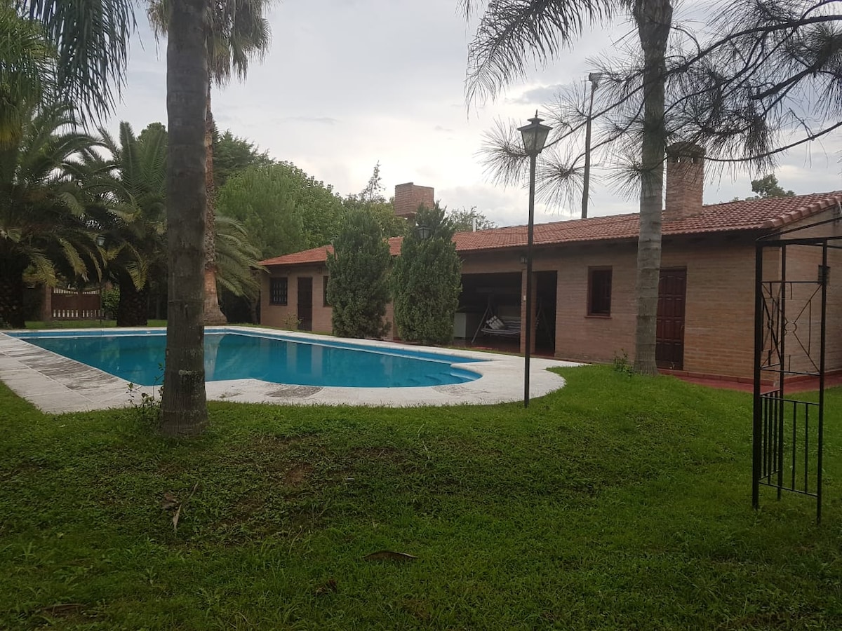 A semi-olympic pool is surrounded by a lush green lawn and palm trees, with lounge chairs positioned nearby. A single-story brick building with a tiled roof is visible in the background, featuring several windows and an outdoor seating area.
