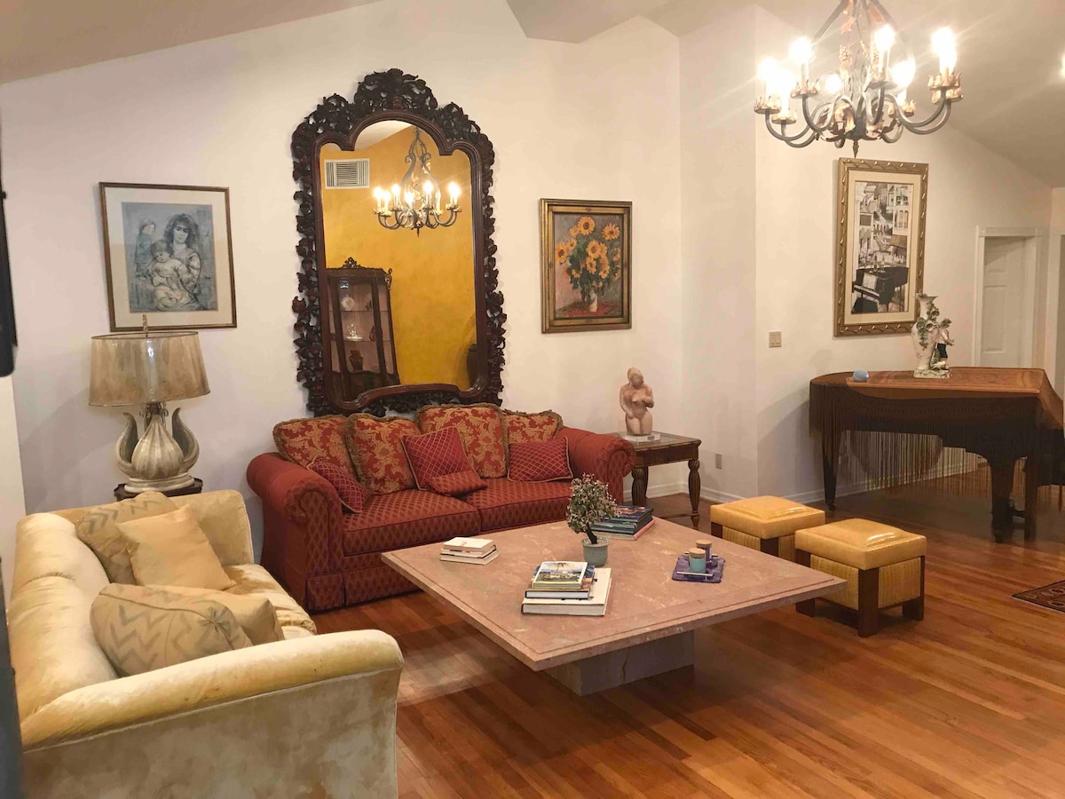 A welcoming living area features a rich color palette with a decorative mirror and artwork adorning the walls. A textured red sofa and a light-toned couch are complemented by a central coffee table and stylish seating cubes. Warm wood flooring enhances the inviting atmosphere.