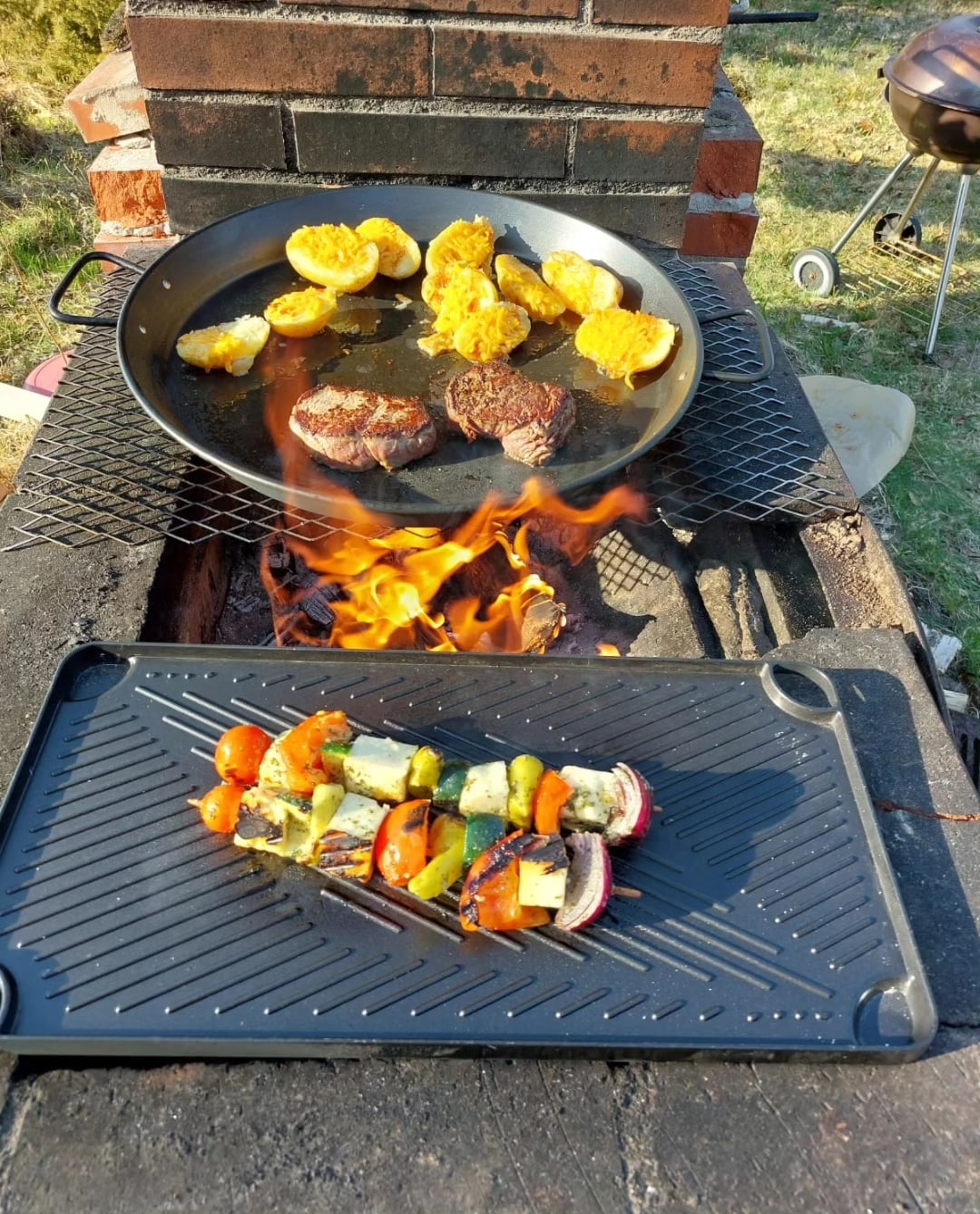 An outdoor cooking area features a grill with various meats, including patties and yellow vegetables, sizzling above an open flame. A tray is positioned beside the flames, displaying colorful skewers of bell peppers and zucchini. The surrounding grass adds a natural touch to the setting.