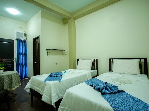 A simple room is furnished with two twin beds dressed in white linens, accented by blue patterned throws. A small table with a floral centerpiece is positioned nearby. A blue curtain covers the window, and air conditioning is mounted on the wall.