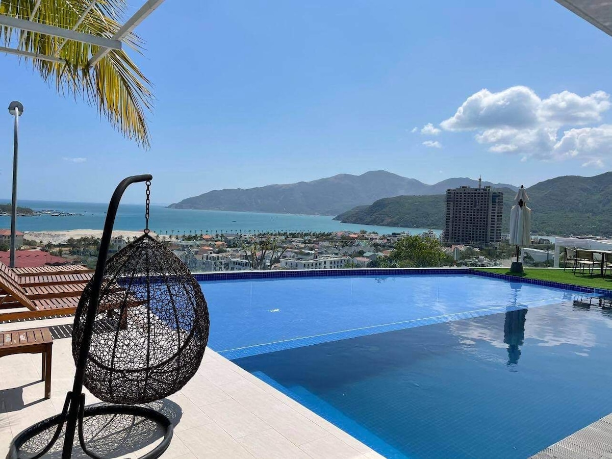 A modern outdoor space features a large infinity pool overlooking a vast ocean and mountainous scenery. A unique hanging chair is positioned beside the pool, with sun loungers nearby. The bright sky complements the tranquil atmosphere, creating a sense of openness and relaxation.