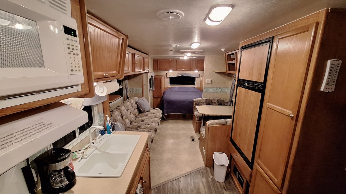 The interior of the camper displays a well-maintained and organized space. A cozy seating area with a patterned couch is positioned beside a compact dining table. A kitchenette is featured on the left, equipped with a sink and microwave. A queen bed is visible at the rear, partially covered by bedding.