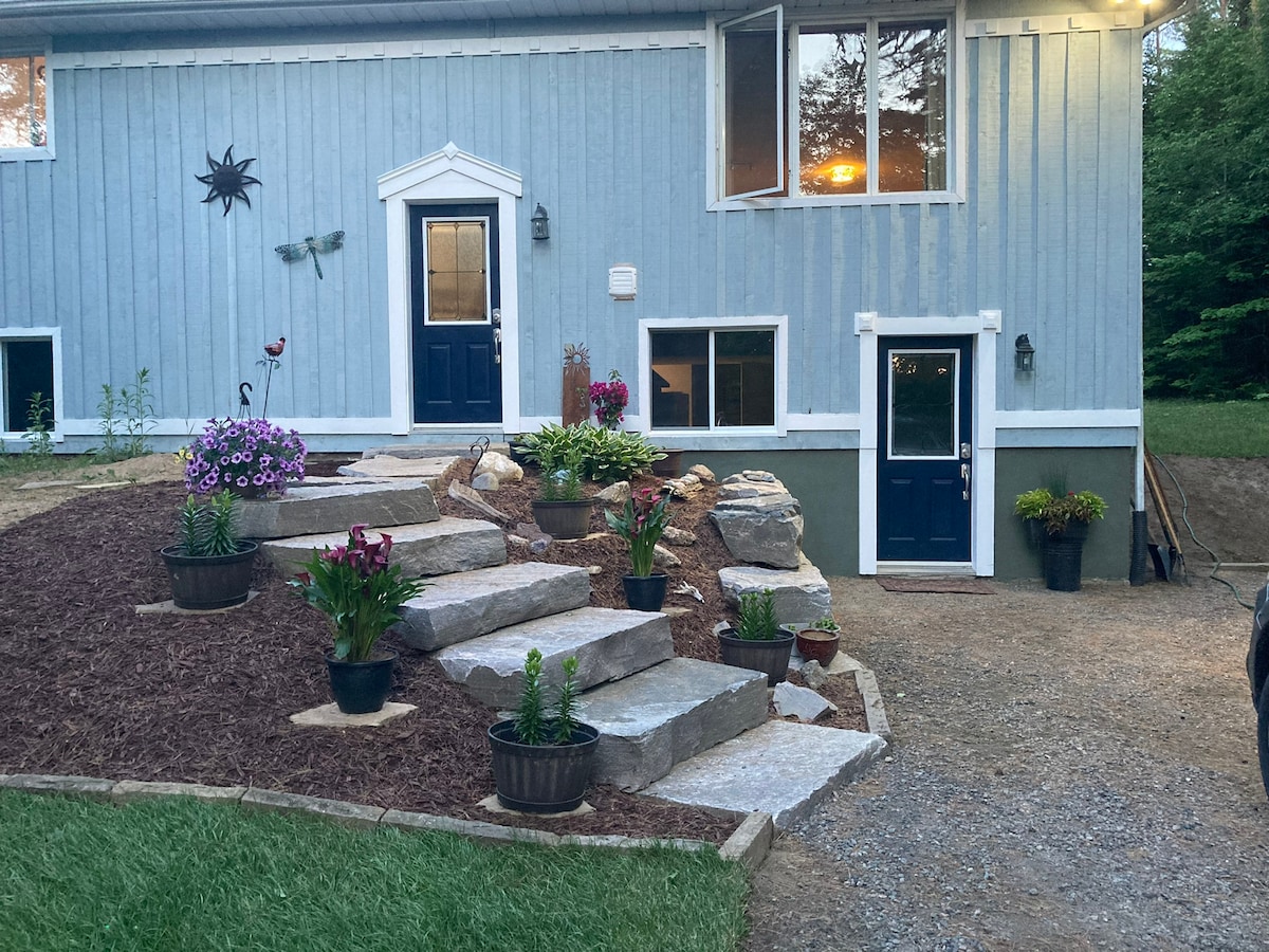 The exterior of a newly renovated lower level apartment is shown, featuring a landscaped path with stone steps leading to the entrance. Colorful flowers in pots add vibrancy, and large windows reflect warm indoor lighting, contributing to the welcoming appearance of the property.