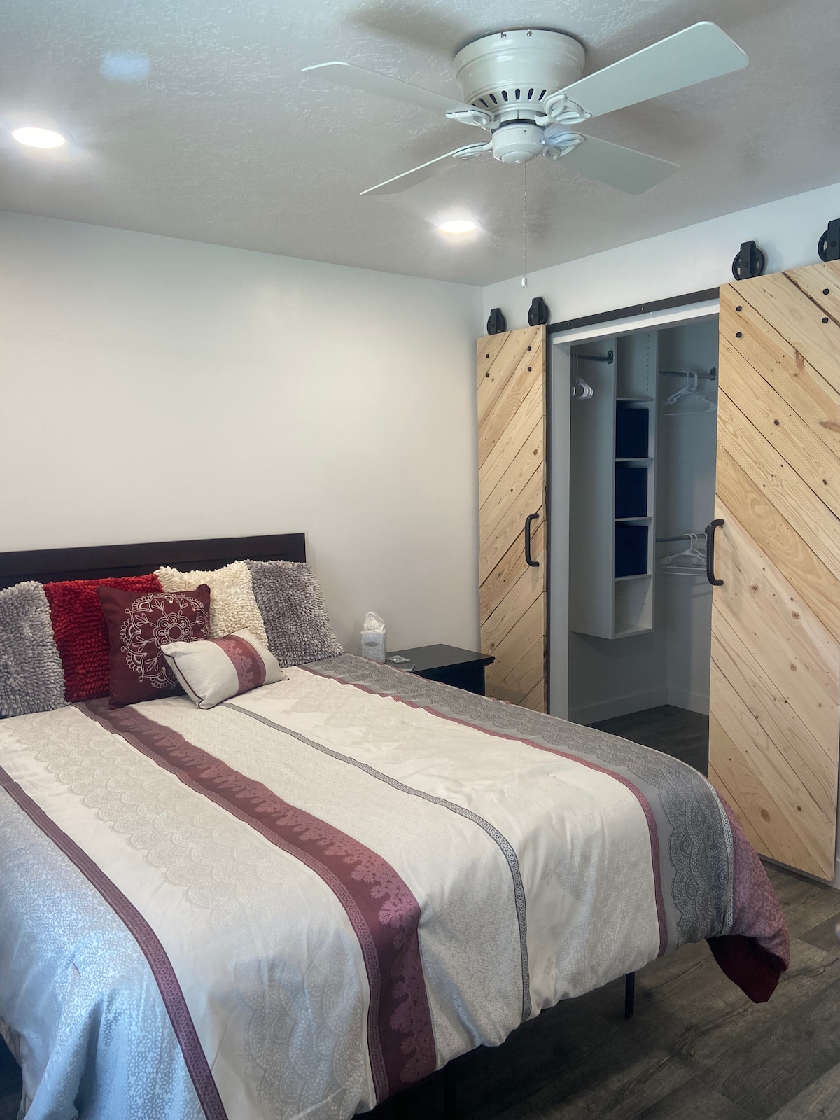 A cozy bedroom features a spacious bed adorned with decorative pillows and a patterned comforter. Natural light is provided by overhead lights, while a ceiling fan enhances airflow. The closet is accessed by wooden sliding doors, revealing clothing storage space.