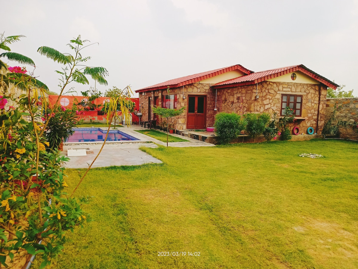 A stone villa is surrounded by a spacious green lawn, with colorful plants and a private pool visible nearby. The red-roofed building features multiple windows and a welcoming entrance, providing a serene and spacious outdoor environment.