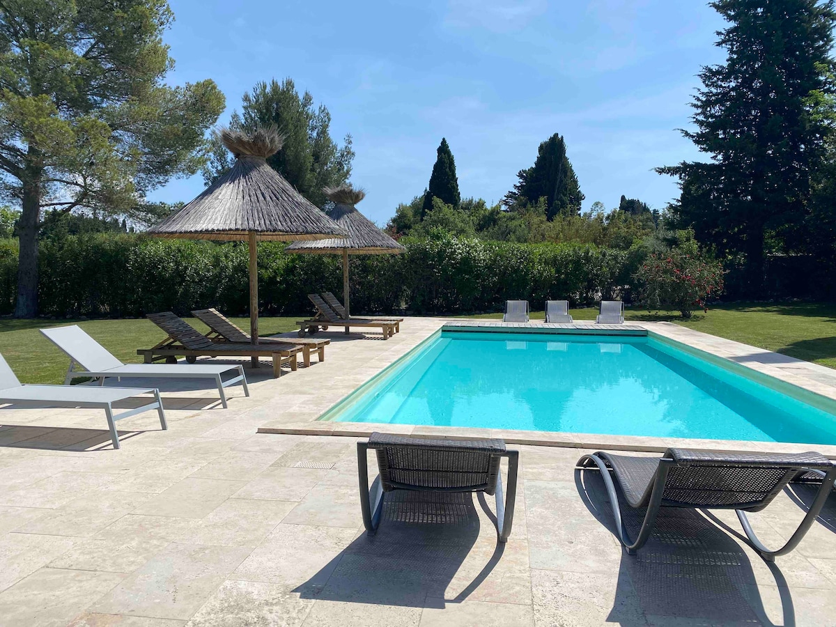 A clear view of a private heated pool is presented, surrounded by well-maintained greenery. Several lounge chairs are arranged on the pool deck, with shade provided by two thatched umbrellas. The pool's inviting turquoise water reflects the sunlight, creating a serene setting.