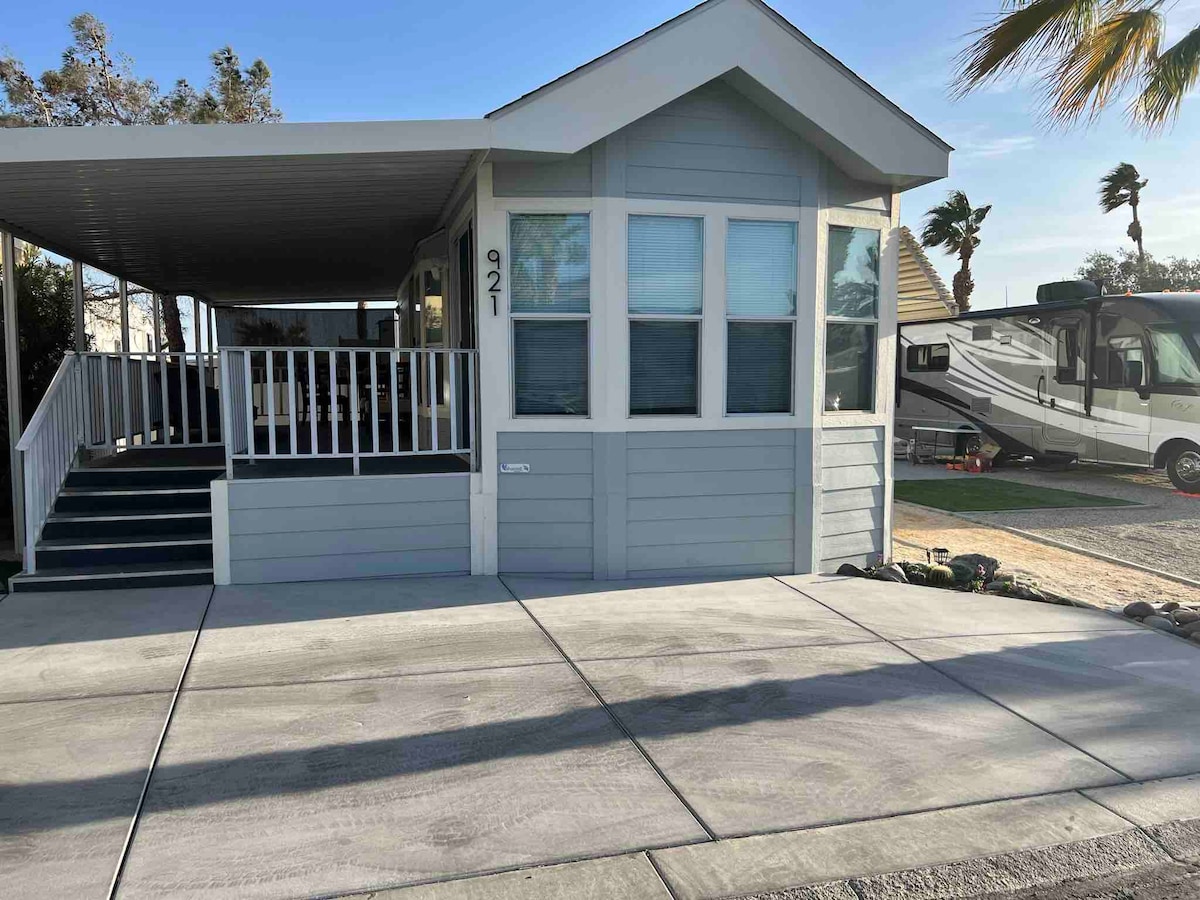 The exterior of the tiny house showcases a light blue facade with white accents, featuring a porch with railings and inviting steps. A concrete driveway leads up to the entrance, with palm trees visible in the background, enhancing the tranquil setting.