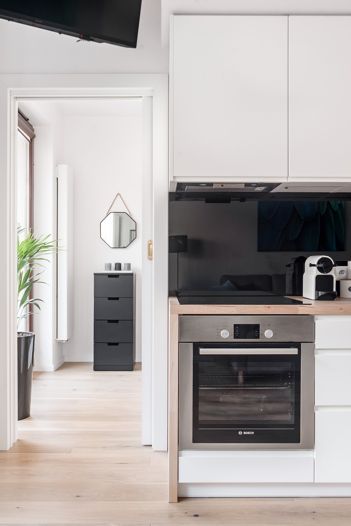 A modern kitchenette is featured, boasting a built-in oven and sleek cabinetry. A tall black dresser is visible through the doorway, complemented by a decorative mirror on the wall. A potted plant adds a touch of greenery, providing a fresh ambiance.