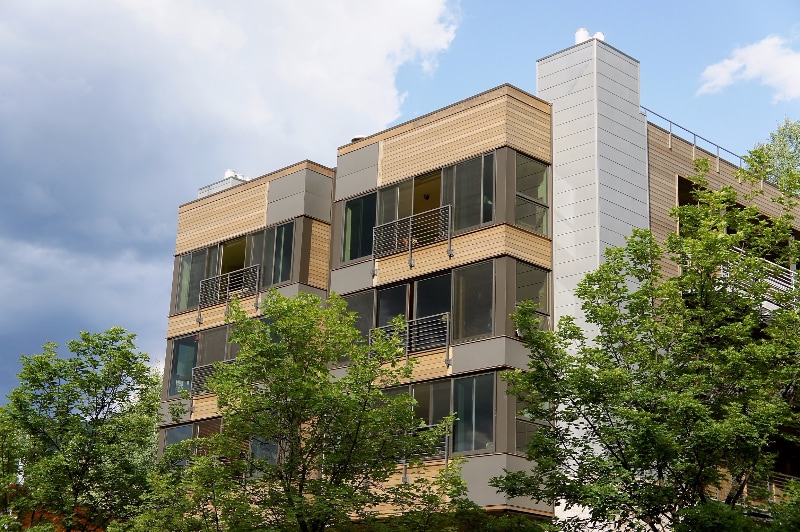 A modern condominium building features a blend of wood and metal exteriors. Expansive windows are framed by greenery, allowing natural light to filter inside. A rooftop terrace is visible, amidst softly clouded skies, enhancing the building's contemporary design.