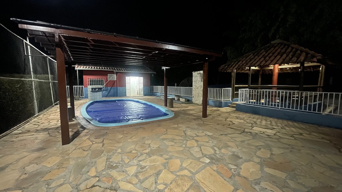 An outdoor area features a covered space adjacent to a heated swimming pool, with a stone patio underfoot. A rustic gazebo is visible in the background, while a basketball hoop is mounted on a wall nearby. Soft lighting enhances the nighttime ambiance.
