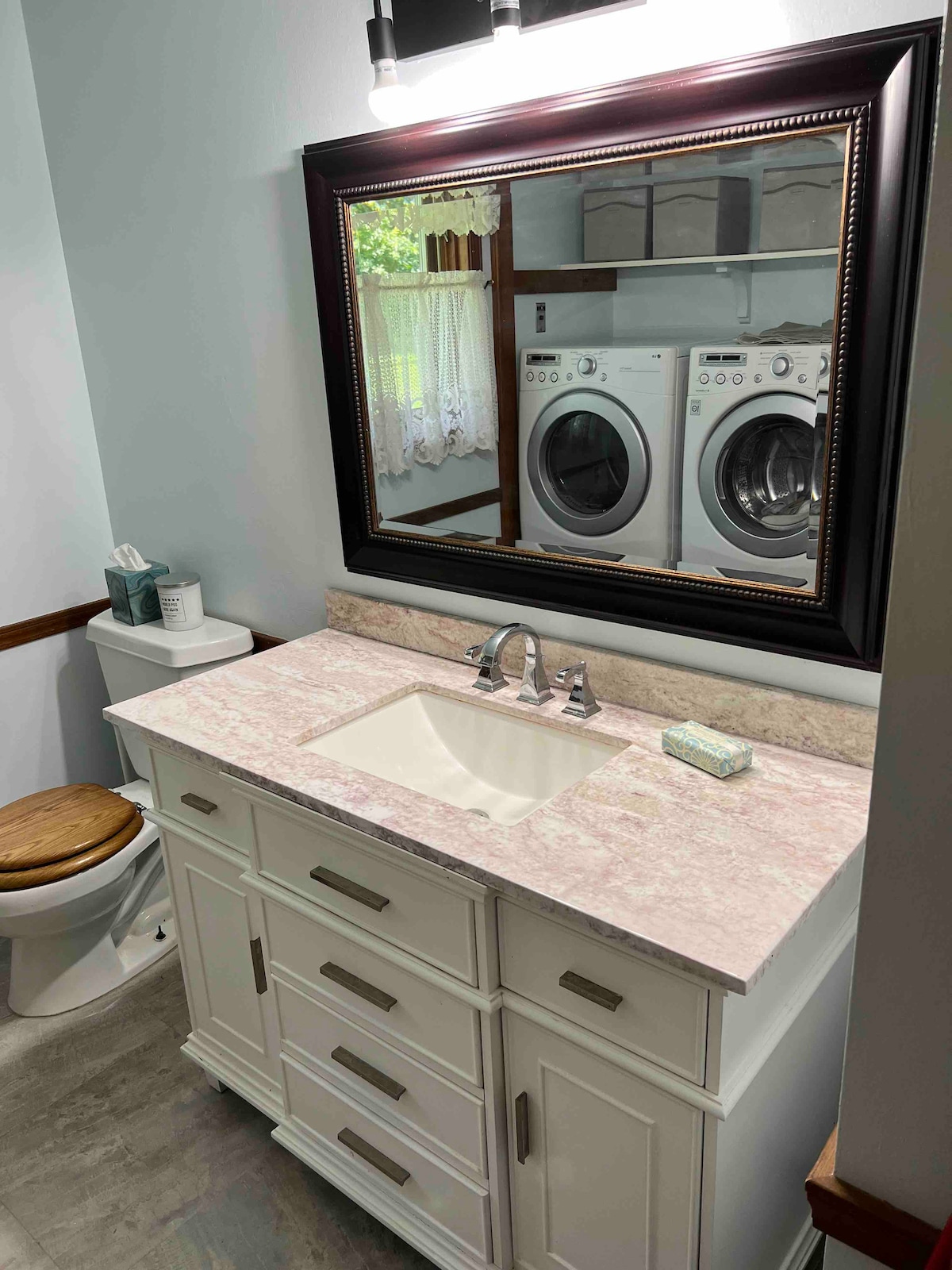 A well-appointed bathroom features a light-colored vanity with a marble countertop and a modern sink. A large mirror reflects the space and the nearby laundry appliances. A wooden stool is positioned next to a clean toilet, while soft natural light is visible through a window draped with lace curtains.