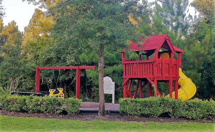 A vibrant red playground is featured, equipped with a slide, swings, and climbing structures. Lush green shrubbery surrounds the play area, providing a natural barrier. Tall trees in the background add shade, while an informational sign stands nearby, indicating the community amenities.