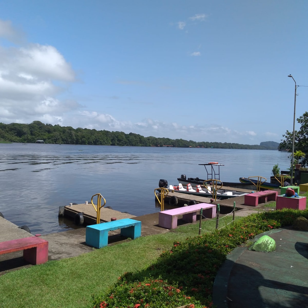 A serene waterway is depicted, showcasing a wooden dock with colorful benches along its edge. Brightly painted structures contrast with the natural greenery in the background, while calm waters reflect the blue sky and distant hills. Trees line the shore, enhancing the tranquil setting.