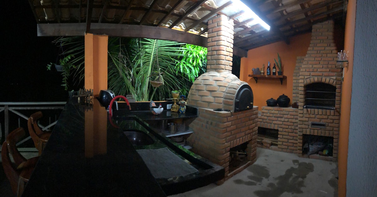 An outdoor gourmet area features a brick pizza oven and two grilling stations. A long dark counter complements the ample space, surrounded by tropical greenery. Soft lighting highlights the textured brick walls, creating a functional cooking environment with an inviting ambiance.