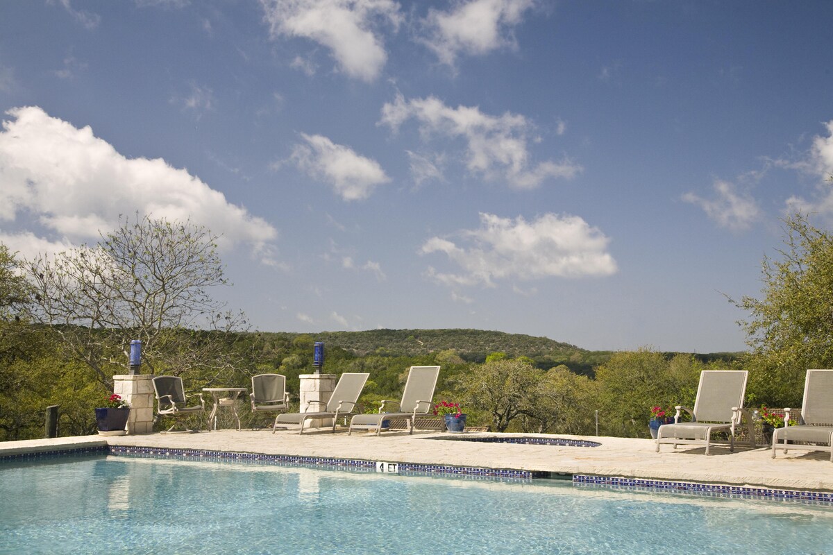 An outdoor pool area is framed by comfortable lounge chairs arranged on a stone patio. The crystal-clear water reflects the sky, while lush greenery and flowering plants enhance the scenic view of the surrounding hills. Soft clouds drift across the blue sky above.