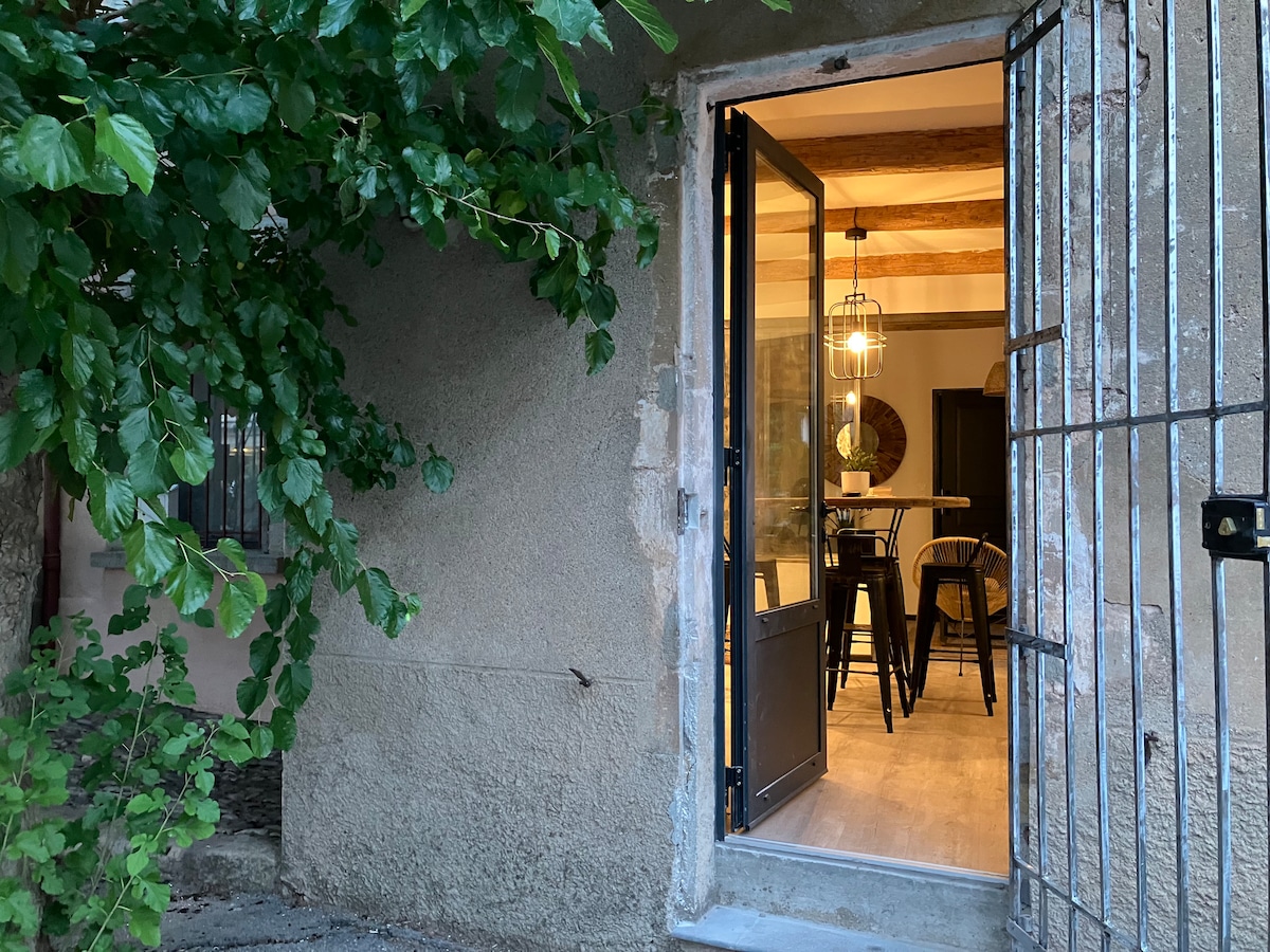 An inviting entrance showcases a black wrought iron gate and a wooden door, partially open, leading into a well-lit interior. Soft light spills from the dining area, highlighting the warm tones of the walls and wooden beams. Lush green foliage frames the entrance, creating a welcoming atmosphere.