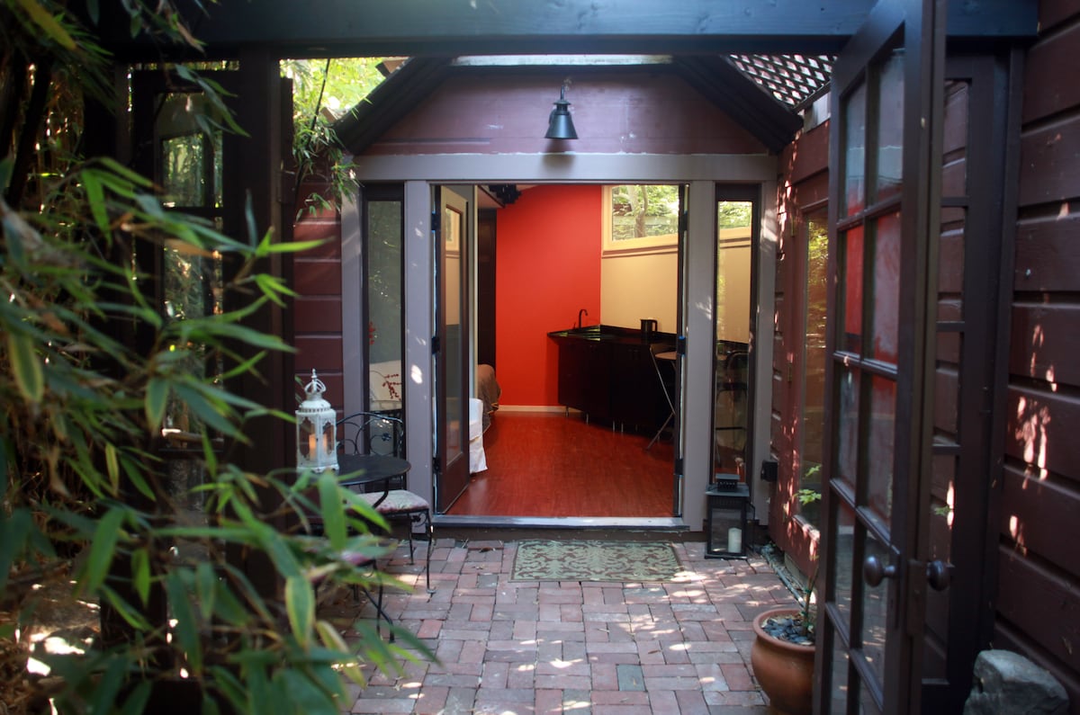 The entrance to the cottage is framed by lush green bamboo. Large glass doors open to reveal a warm interior with red walls and wooden flooring. A small seating area is visible on the brick pathway leading in.