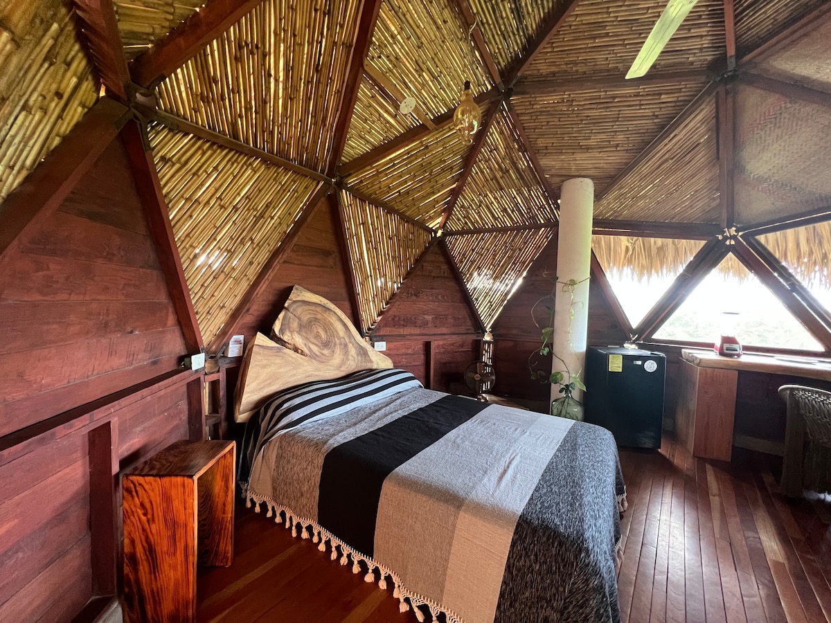 A cozy sleeping area is featured with a spacious bed, adorned with a striped blanket. The room is constructed with natural materials, showcasing bamboo walls and wooden details. Large windows provide views of the surrounding nature, allowing light to fill the space.