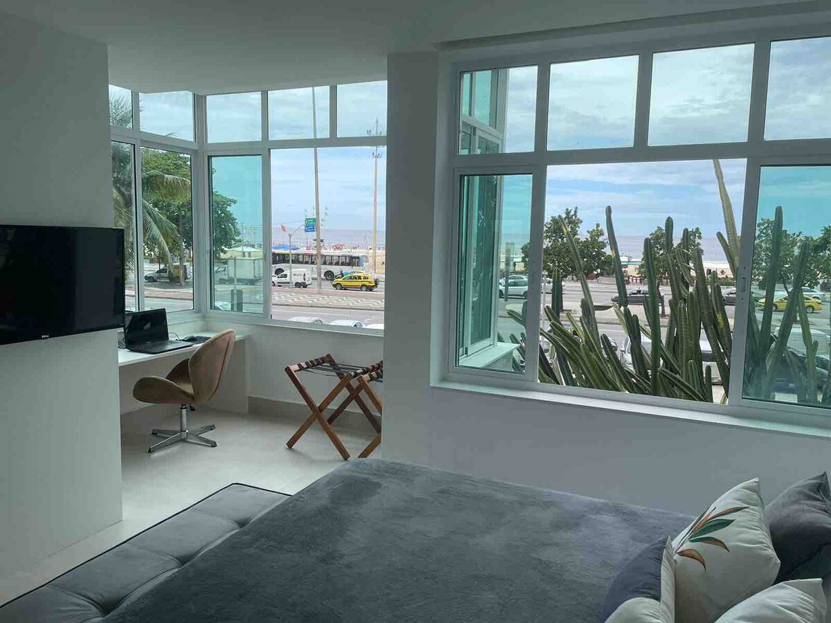 A bright room features large windows offering views of the beach and street. A workspace with a chair is positioned beside a modern television. The bed is dressed with soft linens, complementing the minimalistic decor that enhances the natural light from the windows.