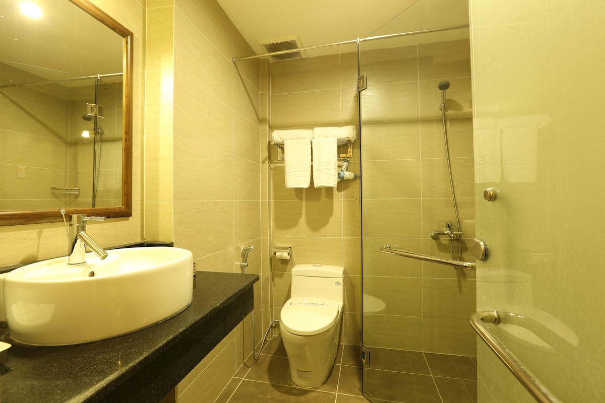 The bathroom features a modern design with neutral-toned tiled walls. A round sink occupies the black countertop, complemented by a large mirror. A glass shower enclosure is visible, along with neatly arranged towels hanging on a rail.